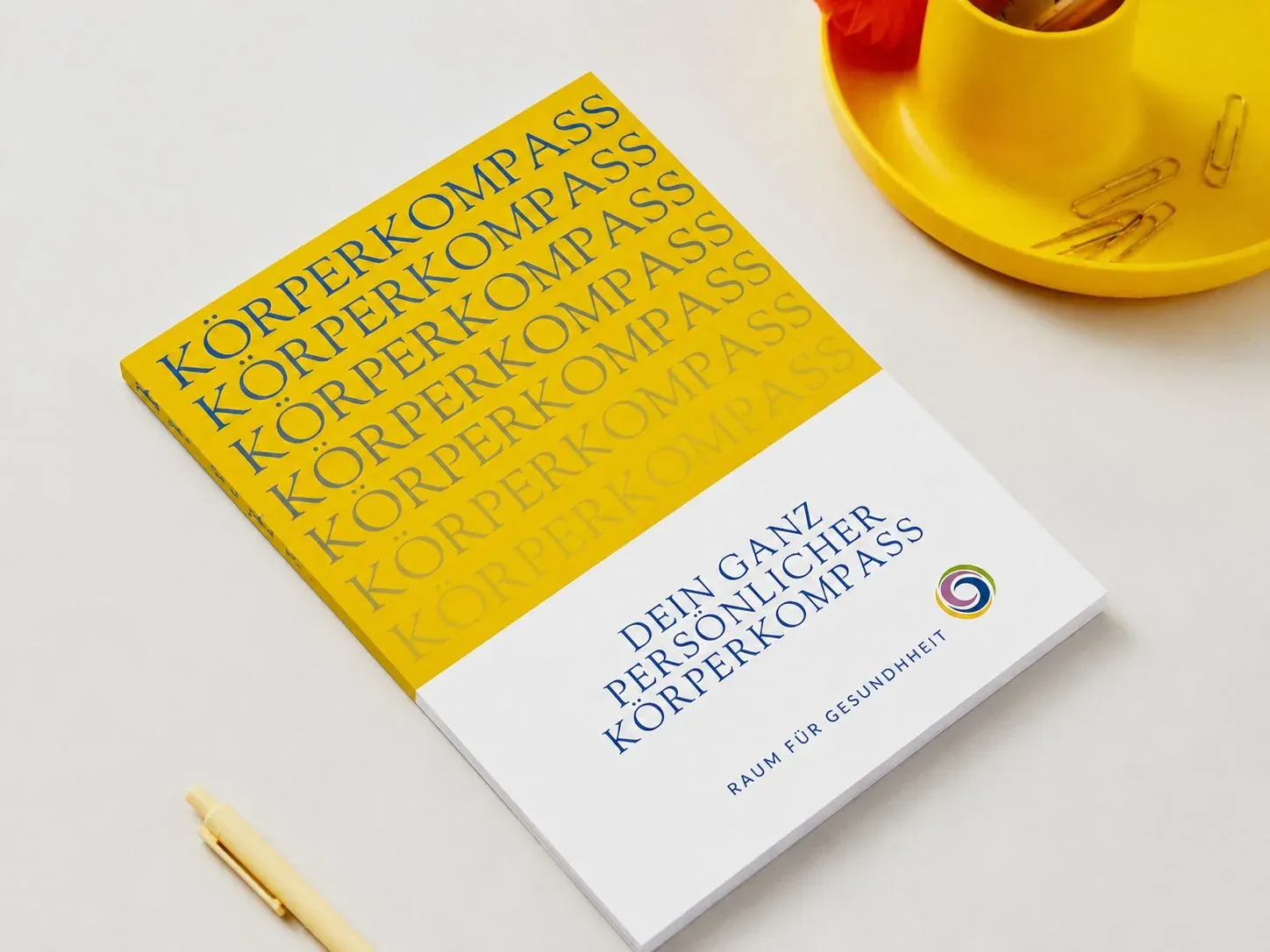 Der Körperkompass als gedrucktes Workbook mit gelbem Umschlag auf einem Schreibtisch